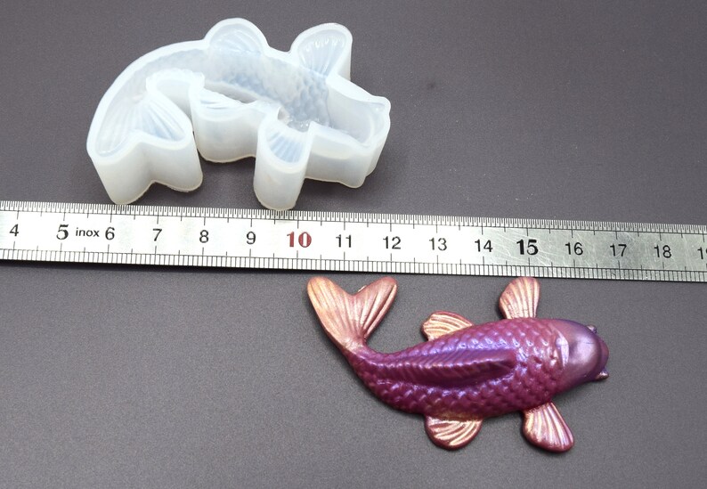 K&ouml;nnte beinhalten: Eine transparente Silikonform eines Koi-Fischs, etwa 10 cm lang, neben einem in Lila und Gold bemalten Koi-Fisch aus Harz.