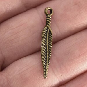 Puede incluir: Un dije de metal dorado en forma de pluma con un dise&ntilde;o detallado. El dije tiene un bucle en la parte superior para sujetarlo a joyas u otras manualidades.