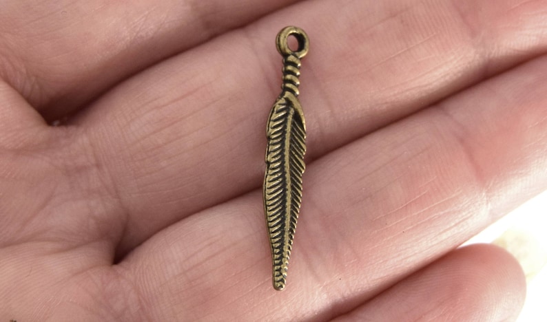 Puede incluir: Un dije de metal dorado en forma de pluma con un dise&ntilde;o detallado. El dije tiene un bucle en la parte superior para sujetarlo a joyas u otras manualidades.