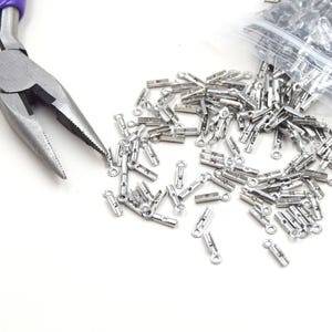 Op de afbeelding: Een close-up van benodigdheden voor het maken van sieraden. Zilverkleurige krimpeindstukken liggen verspreid op een wit oppervlak, met een tang met paarse handvatten in de linkerbovenhoek. Een doorzichtige plastic zak met krimpeinden staat rechtsboven.