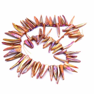 Puede incluir: Un collar de cuentas iridiscentes, de color naranja, con forma puntiaguda y alargada. Las cuentas tienen un efecto brillante multicolor, con toques de púrpura y dorado. Las cuentas están ensartadas en un hilo fino de color claro.