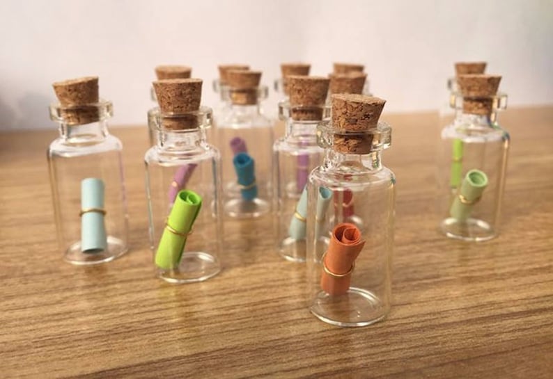May include: A collection of small glass bottles with cork stoppers, each containing a rolled up piece of paper. The bottles are clear and the paper is in various colors.