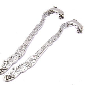 Könnte beinhalten: Zwei silberne Lesezeichen-Anhänger in Form von Delfinen mit detaillierten Designs. Die Delfine sind nach rechts gerichtet und haben oben eine Schlaufe zum Anbringen an einem Lesezeichen.