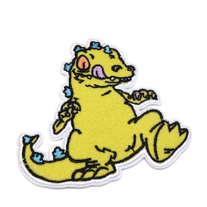 Peut inclure: Dinosaure de dessin animé jaune avec une langue rose qui sort, portant une couronne bleue et violette. Le dinosaure marche avec sa patte droite levée.
