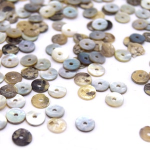 May include: A collection of round, flat, iridescent shell beads in shades of brown, white, and gray. The beads have a hole in the center and are scattered on a white surface.