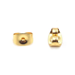 Peut inclure: Deux fermoirs de boucles d'oreilles dorés sont présentés sur fond blanc. L'un est vu de face, avec un petit trou au centre. L'autre est vu de côté, montrant sa forme incurvée.
