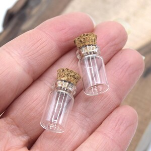 May include: Two small clear glass bottles with cork stoppers. The bottles are about 2 inches tall and are held in a hand.