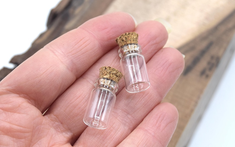 May include: Two small clear glass bottles with cork stoppers. The bottles are about 2 inches tall and are held in a hand.
