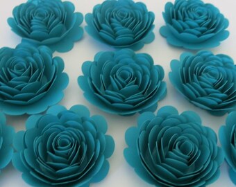 Peut inclure: Dix fleurs en papier turquoise avec des bords festonnés. Les fleurs sont disposées en demi-cercle sur une surface blanche.