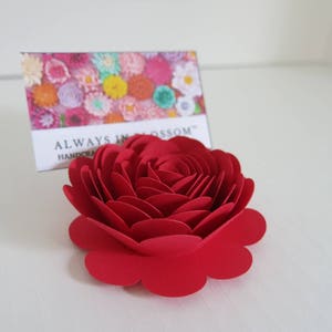 May include: A handcrafted, vibrant red paper rose. The flower is in full bloom, with multiple layers of petals. A card behind the rose reads "ALWAYS IN BLOSSOM HANDCRAFTED". The rose is a decorative item.