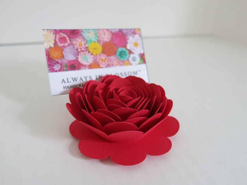 May include: A handcrafted, vibrant red paper rose. The flower is in full bloom, with multiple layers of petals. A card behind the rose reads "ALWAYS IN BLOSSOM HANDCRAFTED". The rose is a decorative item.