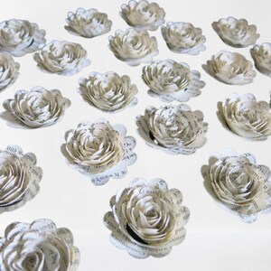 Peut inclure: Une collection de 20 fleurs en papier blanc, chacune faite &agrave; partir de pages d'un livre. Les fleurs sont dispos&eacute;es en rang&eacute;es sur une surface blanche.