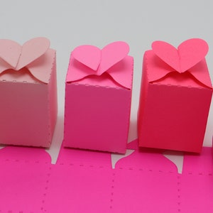 May include: Five pink and red heart-shaped favor boxes with a white coin on top of one of the boxes. The boxes are made of paper and have a dotted pattern.