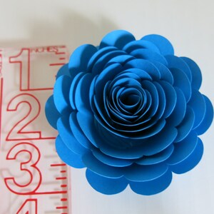 May include: A vibrant blue paper flower, approximately 3 inches in diameter, with layered petals creating a realistic rose appearance. The flower is set against a white background, showcasing its intricate design and color.