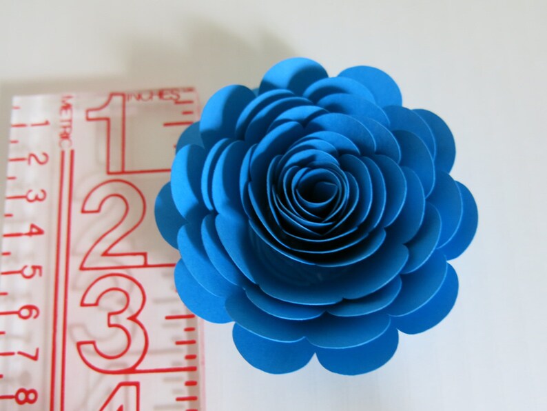 May include: A vibrant blue paper flower, approximately 3 inches in diameter, with layered petals creating a realistic rose appearance. The flower is set against a white background, showcasing its intricate design and color.