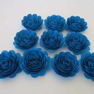 May include: A collection of ten vibrant blue paper flowers arranged on a white surface. Each flower is meticulously crafted with layered petals, creating a realistic rose-like appearance. The flowers are ideal for crafting and decorative projects.