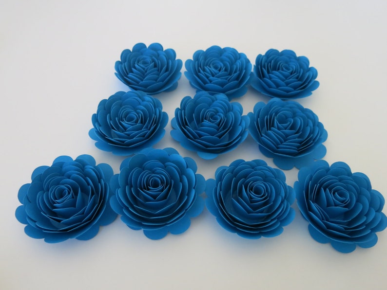 May include: A collection of ten vibrant blue paper flowers arranged on a white surface. Each flower is meticulously crafted with layered petals, creating a realistic rose-like appearance. The flowers are ideal for crafting and decorative projects.