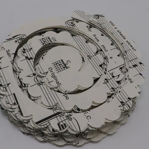 Pode incluir: Uma pilha de c&iacute;rculos de papel branco com bordas festonadas, cada um com uma partitura musical impressa com notas pretas e texto. Um fio de prata &eacute; enrolado em torno da pilha, criando uma forma espiral. O texto na partitura musical diz "Original" e "Just a shade slower".