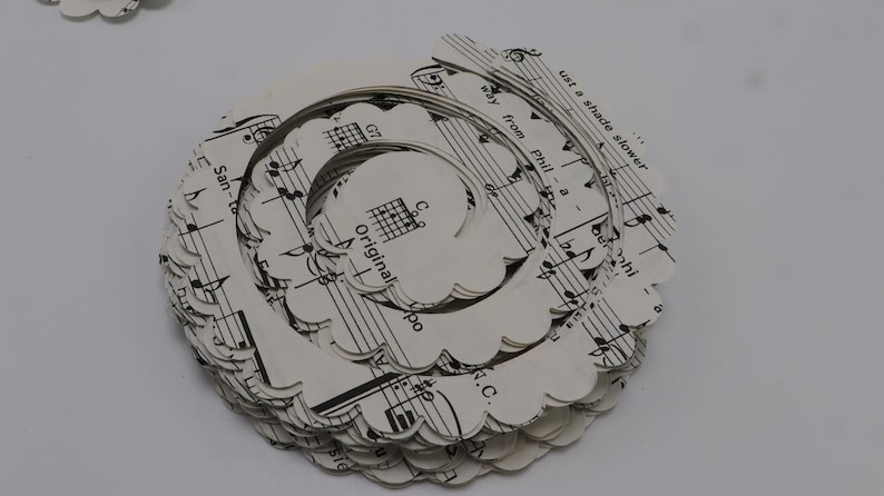 Pode incluir: Uma pilha de c&iacute;rculos de papel branco com bordas festonadas, cada um com uma partitura musical impressa com notas pretas e texto. Um fio de prata &eacute; enrolado em torno da pilha, criando uma forma espiral. O texto na partitura musical diz "Original" e "Just a shade slower".