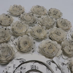 Pode incluir: Uma cole&ccedil;&atilde;o de flores de papel feitas com partituras musicais. As flores s&atilde;o brancas e t&ecirc;m um aspeto delicado e em camadas. As flores est&atilde;o dispostas em um padr&atilde;o circular sobre uma superf&iacute;cie branca.