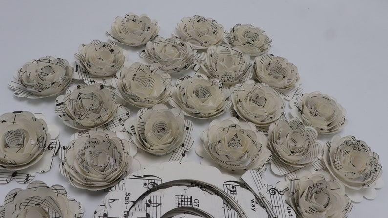 Pode incluir: Uma cole&ccedil;&atilde;o de flores de papel feitas com partituras musicais. As flores s&atilde;o brancas e t&ecirc;m um aspeto delicado e em camadas. As flores est&atilde;o dispostas em um padr&atilde;o circular sobre uma superf&iacute;cie branca.