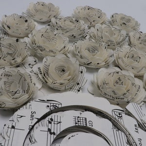 Pode incluir: Uma cole&ccedil;&atilde;o de flores de papel branco feitas com partituras musicais. As flores est&atilde;o dispostas sobre uma superf&iacute;cie branca com mais partituras musicais vis&iacute;veis no fundo.