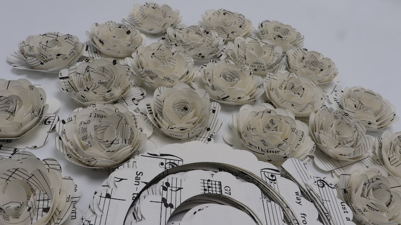 Pode incluir: Uma cole&ccedil;&atilde;o de flores de papel branco feitas com partituras musicais. As flores est&atilde;o dispostas sobre uma superf&iacute;cie branca com mais partituras musicais vis&iacute;veis no fundo.