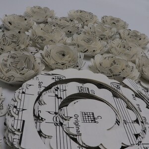 Pode incluir: Uma cole&ccedil;&atilde;o de flores de papel feitas com partituras musicais. As flores s&atilde;o brancas e t&ecirc;m um aspeto delicado e em camadas. As flores est&atilde;o dispostas em c&iacute;rculo em torno de uma folha de m&uacute;sica maior com a palavra "Original" impressa nela.
