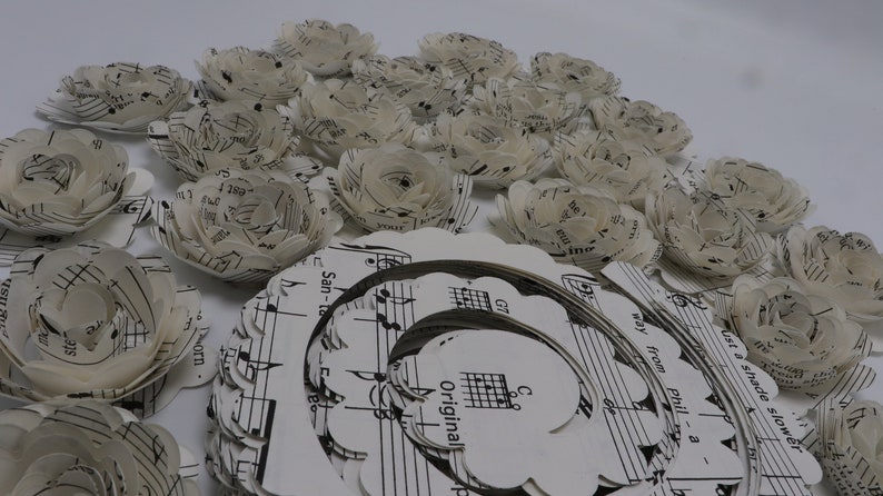 Pode incluir: Uma cole&ccedil;&atilde;o de flores de papel feitas com partituras musicais. As flores s&atilde;o brancas e t&ecirc;m um aspeto delicado e em camadas. As flores est&atilde;o dispostas em c&iacute;rculo em torno de uma folha de m&uacute;sica maior com a palavra "Original" impressa nela.