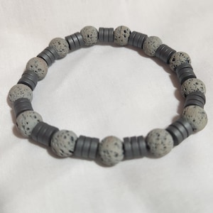 May include: A gray bracelet featuring a combination of round, speckled beads and flat, cylindrical beads. The bracelet is made of a dark gray material and is displayed on a white surface. The beads are evenly spaced, creating a simple, yet stylish accessory.