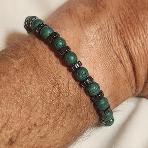 Puede incluir: Una pulsera de cuentas verdes con detalles metálicos de color gris oscuro. La pulsera presenta cuentas verdes redondas y texturizadas intercaladas con pequeñas piezas metálicas rectangulares. La pulsera se lleva en la muñeca.