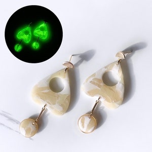 Peut inclure: Une paire de boucles d'oreilles crème avec un motif marbré et des accents dorés. Les boucles d'oreilles ont une forme unique avec une découpe circulaire et un charm circulaire pendant. Une version verte lumineuse est montrée en haut à gauche.