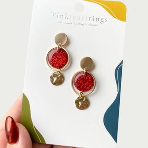 May include: Gold-toned earrings with red glitter-filled circles and hammered metal accents. The earrings are displayed on a white card with the text "Tink[ear]rings" and "Handmade by Maggie Michel".