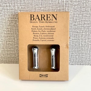 Peut inclure: Un paquet de deux boutons de porte chromés, étiqueté "BAREN DESIGN: TORD BJÖRKLUND" avec le logo IKEA. L'emballage comprend plusieurs descriptions linguistiques pour les boutons, notamment "Knopp, 2-pack, förkromád", "Knob, 2-pack, chrome-plated", "Haken, 2er Pack, verchromt", "Bouton, 2 pièces, chromé", "Knop, 2 st, verchroomd", "Pomo, 2 piezas, cromado", et "Pomello, 2 pezzi, cromato".