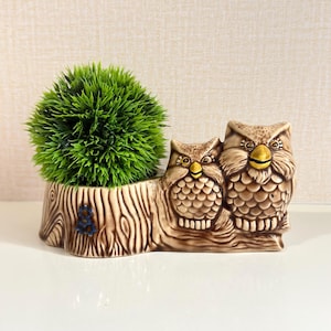 May include: A ceramic planter shaped like a tree log with two brown owls perched on the side. The owls have yellow beaks and detailed feather textures. A small blue flower is moulded onto the log. A round, vibrant green artificial plant sits inside.