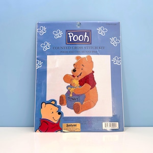 May include: A blue counted cross stitch kit featuring Winnie the Pooh. The kit includes an image of Pooh with a honey pot, and the text "Pooh and the Hunny Pot". The kit also includes the text "Janlynn".