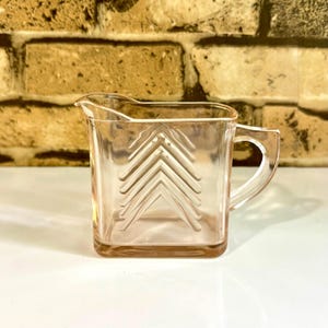 May include: A vintage, pink glass creamer with a square base and a handle. The creamer features a series of embossed chevrons on the front. The creamer is approximately 4 inches tall and is set against a brick background.