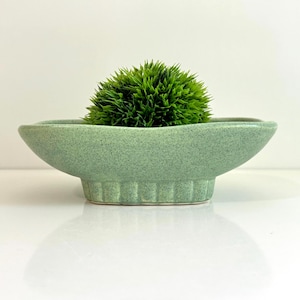 May include: A light green speckled ceramic bowl with a shallow, elongated oval shape and a fluted base. The bowl contains a dense, rounded, bright green artificial plant with spiky foliage that sits centred within the vessel.