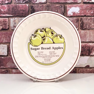 May include: A round, off-white ceramic pie plate with a brown rim and a decorative apple graphic. The plate features the text "Sugar Bread Apples" and a recipe. The plate is displayed on a clear stand against a brick background.