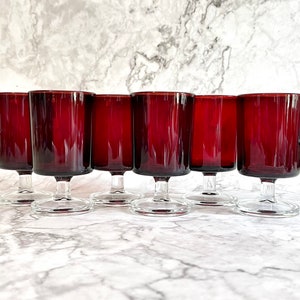 May include: A set of six vintage wine glasses. Each glass has a deep red, rectangular bowl and a clear, stemmed base. The glasses are arranged in a row, highlighting their consistent design and colour. The glassware is suitable for serving wine.