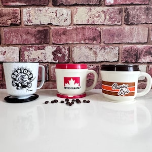 May include: Three vintage coffee mugs with handles. One mug features a Raptors logo, another has a Petro-Canada logo, and the third has a "Country Style" design. The mugs are white with red and brown accents, and are displayed on a white surface.
