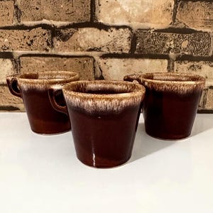 May include: Three brown ceramic mugs with a dark brown exterior and a lighter brown and white rim. Each mug has a handle and a glossy finish. The mugs are arranged on a white surface against a brick wall.