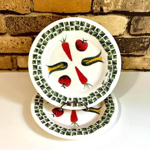 May include: Two white ceramic plates with a green mosaic border and painted vegetables. The plates feature red tomatoes, carrots, and yellow squash. A metal stand holds the bottom plate.