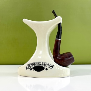 May include: White ceramic pipe stand with the words "Barber's Saloon" in black lettering. A brown wooden pipe is resting in the stand.