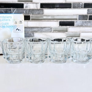 May include: A set of clear glass candle holders with a faceted design. The candle holders are arranged in a row on a white surface, with a box in the background that reads "Chandeliers, Coquetiers, Egg cups, Candle holders."