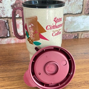 May include: A beige travel mug with a black lid and a red handle. The mug has a graphic of a cartoon character and the text "Saint Cinnamon Premium Blend Coffee". The mug has a matching red lid with a push button.