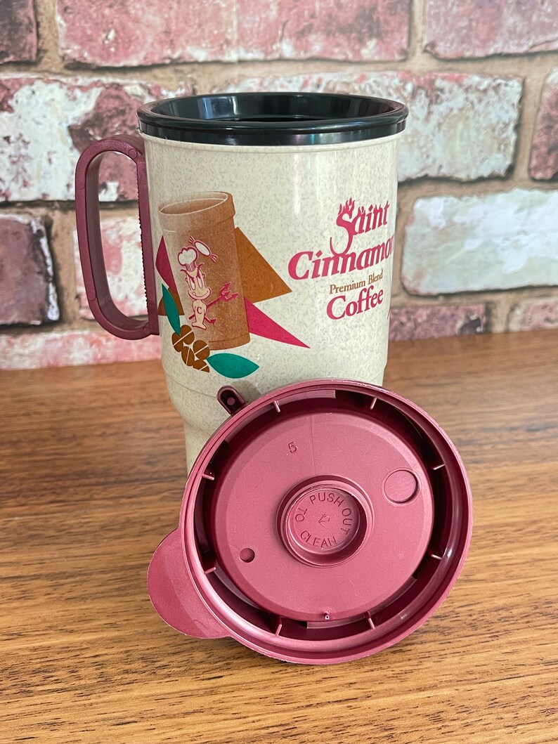 May include: A beige travel mug with a black lid and a red handle. The mug has a graphic of a cartoon character and the text "Saint Cinnamon Premium Blend Coffee". The mug has a matching red lid with a push button.