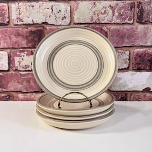 May include: A set of four off-white ceramic plates with a concentric spiral design. Each plate has a gray band around the center and a speckled rim. The plates are stacked on a gold-colored metal stand against a brick wall background.