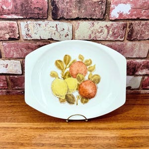 May include: White oval serving dish with a raised rim and a citrus fruit design. The design features three fruits: two yellow lemons and one orange. The fruits are surrounded by green leaves.