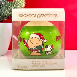 May include: A green Christmas ornament with a cartoon character wearing a red Santa hat and a green scarf. The ornament also features the text "Seasons Greetings" and "1979".